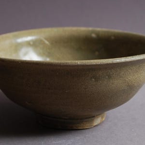 Könnte beinhalten: Eine Keramikschale mit abgerundeter Form und hellbrauner Glasur. Die Schale hat einen leicht ausgestellten Rand und einen kleinen, kreisförmigen Fuß. Die Oberfläche hat ein strukturiertes Aussehen, mit kleinen sichtbaren Unvollkommenheiten. Die Schale ist wahrscheinlich zum Servieren von Speisen.