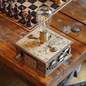 Op de afbeelding: Een vierkante houten tafellamp met een heldere Edison-stijl gloeilamp gemonteerd op een messing fitting. Het bovenoppervlak toont gedrukte technische schema's, een ronde draaiknop en een tuimelschakelaar. De zijpanelen bevatten kleine zwarte schakelaars.