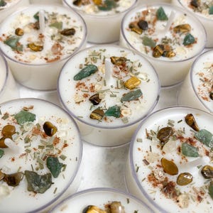 May include: A collection of white tea light candles in clear plastic cups, each topped with small green and brown polished stones, dried herbs, and a dusting of reddish-brown powder, featuring a central white wick protruding from the wax surface.