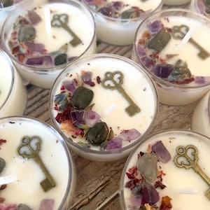 May include: A collection of round tea light candles in clear plastic containers. Each white wax candle is topped with a bronze-colored metal key charm, small purple amethyst crystals, dark green stones, and dried flower petals around a central wick.