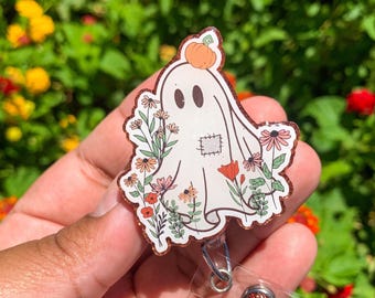 May include: A badge reel featuring a white ghost illustration with a small orange pumpkin on its head and a patch on its chest. The ghost is surrounded by colorful wildflowers in shades of pink, orange, and green, with a glittery copper-toned border.