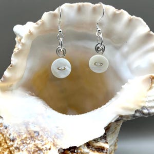 May include: Button earrings with silver hooks. Each earring has a small, round, off-white button with a curved slot. The earrings are presented inside a large, white seashell.