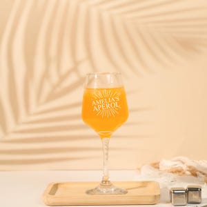 May include: A clear stemmed glass filled with an orange beverage, with the text "AMELIA'S APEROL" in white. The glass sits on a wooden tray. Two stainless steel ice cubes are next to the tray. The background is a light beige color.