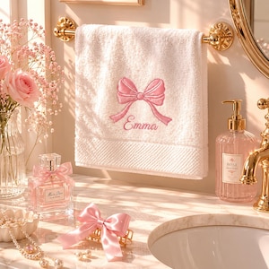 May include: A white hand towel featuring an embroidered pink bow and the name Emma in pink script, hanging on a gold towel bar. The towel has a textured border at the bottom and is displayed in a bathroom setting with various vanity accessories.