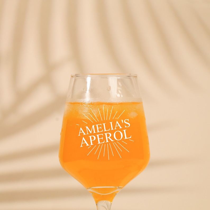 May include: A clear wine glass filled with an orange beverage. The glass has the text "AMELIA'S APEROL" in white. The background is a light tan color with a palm leaf shadow.