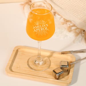 May include: A stemmed glass filled with an orange beverage, resting on a wooden tray. The glass has the text "AMELIA'S APEROL" printed on it. Stainless steel ice cubes and tongs are also on the tray.