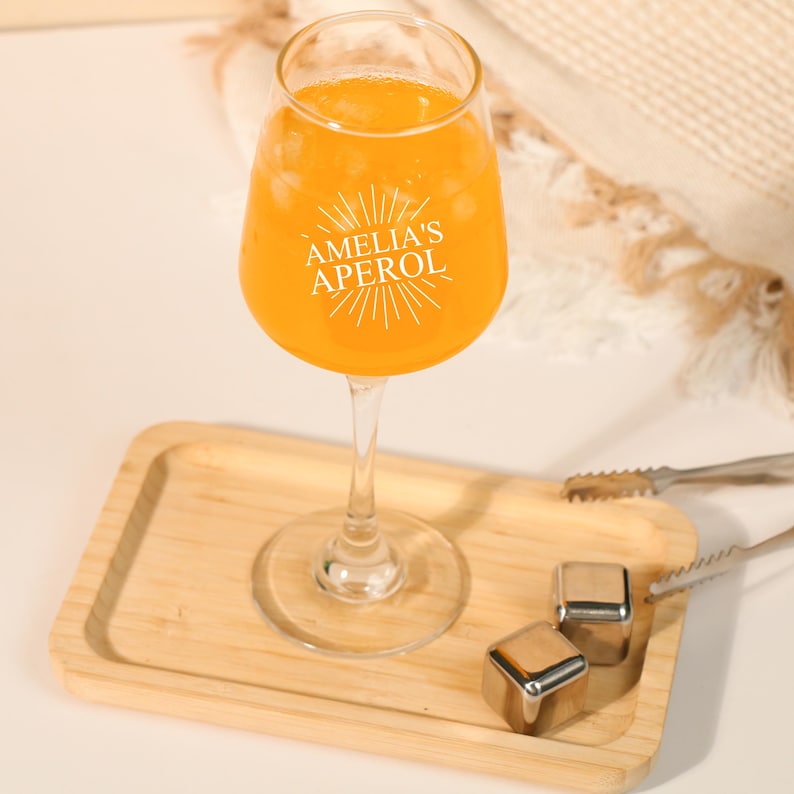 May include: A stemmed glass filled with an orange beverage, resting on a wooden tray. The glass has the text "AMELIA'S APEROL" printed on it. Stainless steel ice cubes and tongs are also on the tray.