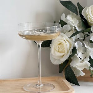 Könnte beinhalten: Ein klares Champagnerglas mit dem eingravierten Namen "Isabella" auf der Seite, gefüllt mit einer hellen Flüssigkeit. Das Glas steht auf einem Holztablett, mit weißen Rosen und Grünzeug im Hintergrund. Perfekt für besondere Anlässe.