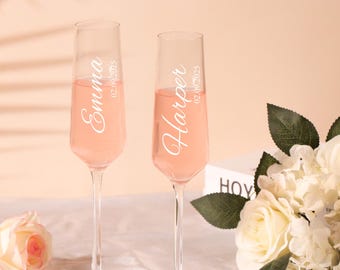May include: Two champagne flutes filled with pink liquid, personalised with the names "Emma" and "Harper" in elegant white script. The date "02.09.2025" is printed below each name. The flutes are set on a table with flowers.