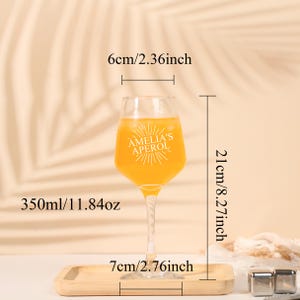 May include: A clear wine glass filled with an orange liquid, labeled "AMELIA'S APEROL". The glass is 8.27 inches tall, 2.36 inches wide, and holds 11.84 oz. It sits on a wooden tray that is 2.76 inches wide.