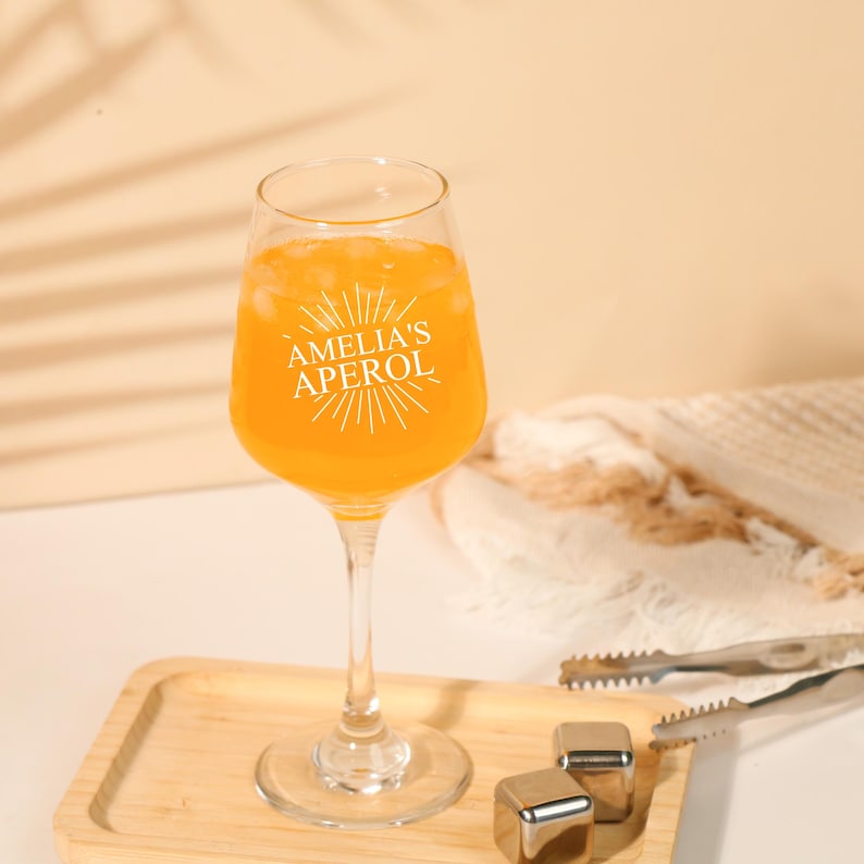 May include: A clear wine glass filled with an orange beverage and ice. The glass has the text "AMELIA'S APEROL" in white. The glass sits on a wooden tray with stainless steel ice cubes and tongs. The background is a neutral color.