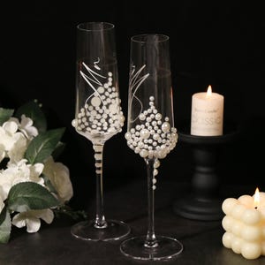 May include: Two champagne flutes decorated with white pearl-like beads and the words "Mr." and "Mrs." written in white on the glass. The flutes are on a black surface with two white candles.