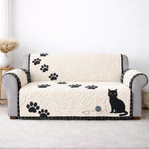 May include: Cream-colored sofa cover with black paw prints and a black cat design. The cover features a quilted pattern and a black cat sitting next to a ball of yarn. The edges are trimmed with a plaid pattern.