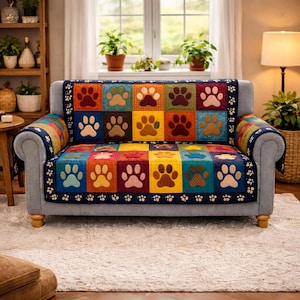 Puede incluir: Un sofá gris de dos plazas con un edredón colorido con huellas de patas. El edredón presenta un diseño de patchwork con cuadrados de varios colores, cada uno mostrando una huella de pata. Los bordes del edredón están ribeteados con tela azul oscuro y huellas de patas blancas.