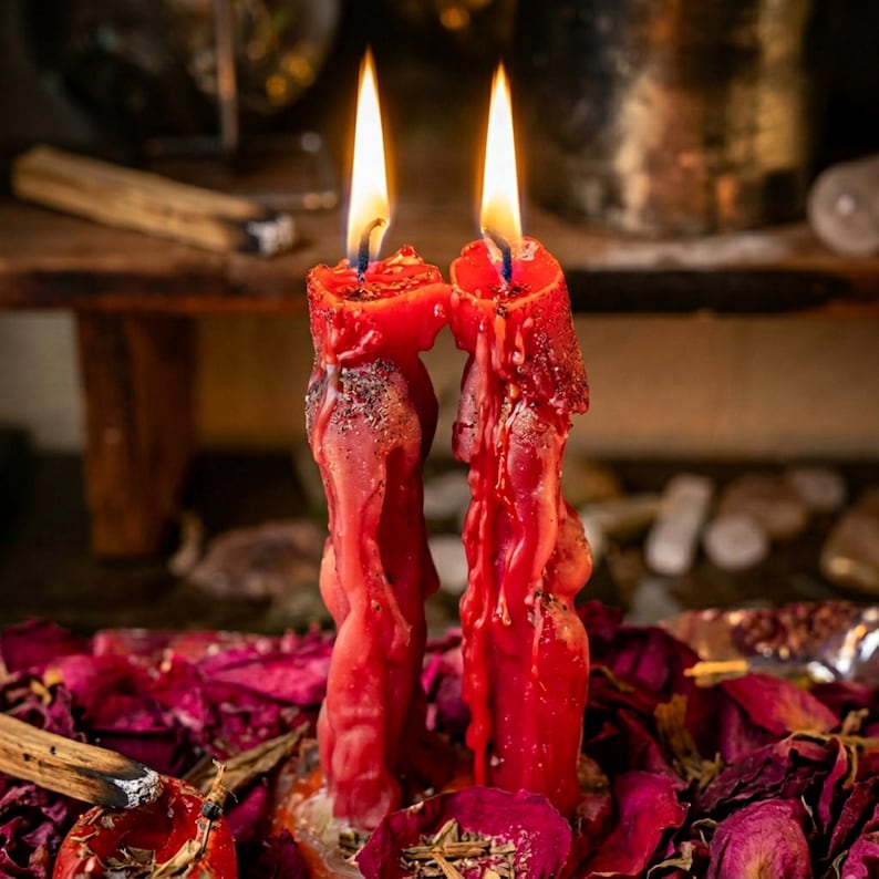 May include: Two red, lit candles with dripping wax, set against a backdrop of dried rose petals and a wooden surface. The candles have a textured appearance and are surrounded by a mystical ambiance.