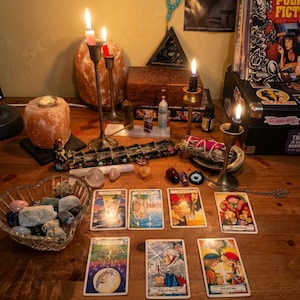 May include: A wooden table displays a spiritual setup with lit candles in brass holders, tarot cards, crystals, and a heart-shaped bowl of stones. A wooden box, incense, and a vintage record player are also present.
