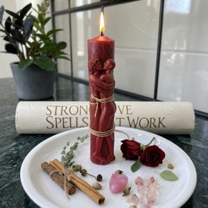 May include: A lit, burgundy-colored candle with a carved design of embracing figures, tied with twine. The candle sits on a white plate with cinnamon sticks, rose buds, and crystals. A rolled scroll behind the candle reads "STRONG SPELLS LOVE THAT WORK."