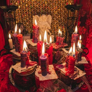 May include: A collection of lit candles in various colors, including red, purple, and white, arranged on glass jars and a red surface. A white angel statue is in the background, with a gold mesh backdrop. The candles are of different sizes and shapes.