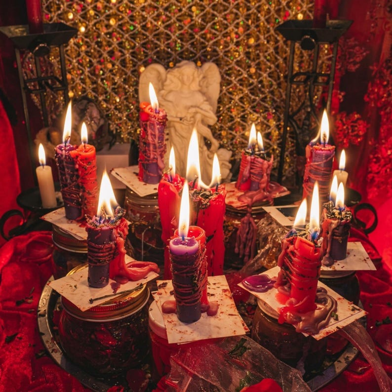 May include: A collection of lit candles in various colors, including red, purple, and white, arranged on glass jars and a red surface. A white angel statue is in the background, with a gold mesh backdrop. The candles are of different sizes and shapes.