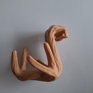 Puede incluir: Un gancho de pared de cerámica de color marrón claro, con forma de mano. El gancho tiene forma de mano con los dedos curvados hacia adentro.