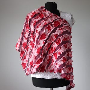 May include: A red and pink textile shawl with a floral pattern. The shawl is draped over a white top on a black mannequin. The design features layers of ruffled fabric in shades of red and pink, creating a textured and visually appealing accessory.