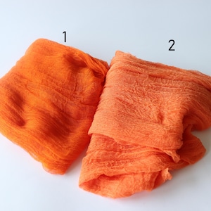 Op de afbeelding: Twee stukken feloranje stof worden getoond tegen een witte achtergrond. De stof heeft een gekreukte textuur en de stukken zijn genummerd met de nummers 1 en 2. De stof lijkt licht van gewicht en kan worden gebruikt voor knutselen of decoratie.