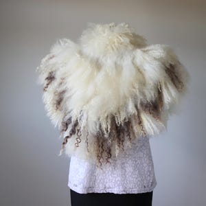 Könnte beinhalten: Ein flauschiger, cremefarbener und brauner Schal mit einem strukturierten, lockigen Design. Der Schal ist über einem weißen Spitzentop und einem schwarzen Unterteil drapiert. Der Schal scheint aus einem weichen, natürlichen Material zu bestehen.
