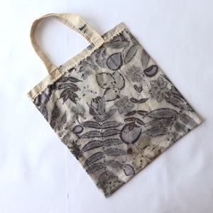 Puede incluir: Una bolsa de tela beige con un estampado floral en tonos grises y marrones. La bolsa tiene una textura de tela natural y un dise&ntilde;o sencillo.