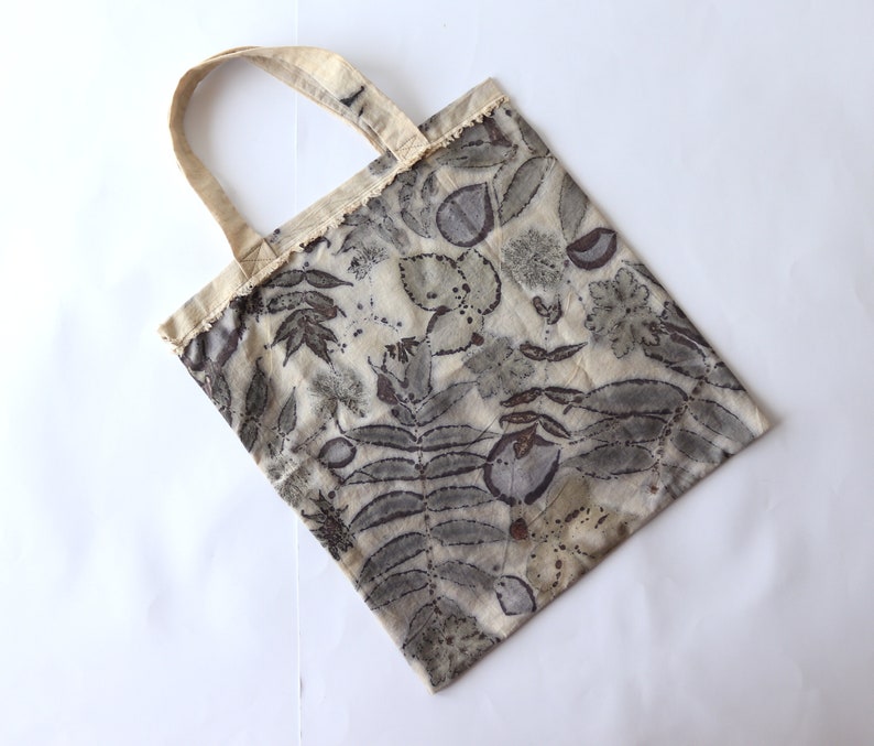 Puede incluir: Una bolsa de tela beige con un estampado floral en tonos grises y marrones. La bolsa tiene una textura de tela natural y un dise&ntilde;o sencillo.