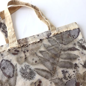 Puede incluir: Una bolsa de tela beige con estampados de hojas marrones y manchas marrones. La bolsa tiene un borde deshilachado y un asa de tela beige.