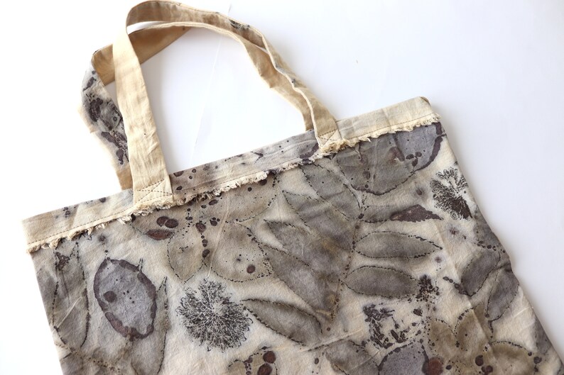 Puede incluir: Una bolsa de tela beige con estampados de hojas marrones y manchas marrones. La bolsa tiene un borde deshilachado y un asa de tela beige.