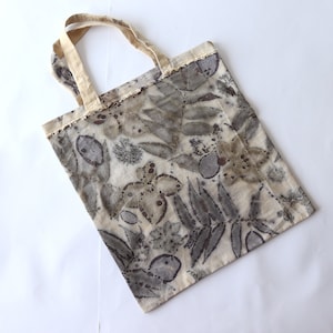 Puede incluir: Una bolsa de tela beige con un estampado de hojas y flores en tela natural en tonos marrones y grises. La bolsa tiene un asa de tela natural.