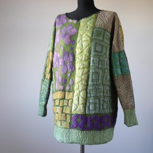 May include: A patchwork sweater in shades of green, purple, and tan. The sweater features a variety of textures and patterns, including floral, geometric, and brick-like designs. The long-sleeved sweater is displayed on a black mannequin.