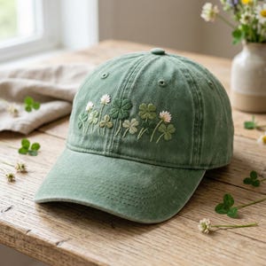 May include: A green baseball cap featuring an embroidered design of clovers and small white flowers. The cap is made of a soft, worn fabric and has a curved brim. The embroidery features varying shades of green and white.