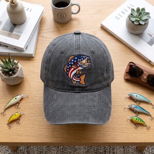 May include: A charcoal grey baseball cap featuring an embroidered graphic of a fish with a red, white, and blue American flag pattern on its body. The cap has a distressed, washed-out appearance with a curved brim and a button on the top crown.