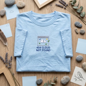 May include: A light blue t-shirt featuring an embroidered graphic of a computer window containing a broken cloud, a sun, a bird, and leaves. Below the graphic, the text reads 404 CLOUD NOT FOUND in dark blue, capital, sans-serif letters.