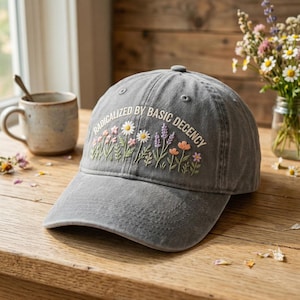 May include: Grey baseball cap with embroidered floral design and the text "RADICALIZED BY BASIC DECENCY". The cap has a curved peak and a button on top. The embroidery includes daisies, lavender, and other colourful flowers.