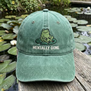 May include: Green baseball cap with a curved brim. The cap features an embroidered design of a frog on a lily pad, with the text "MENTALLY GONE" below the frog. The cap is made of a soft, worn material.
