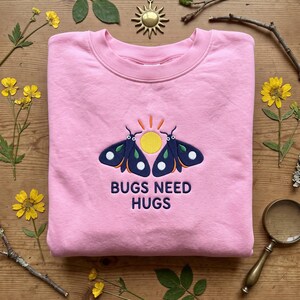 May include: A light pink crewneck sweatshirt featuring an embroidered design of two dark blue moths flanking a yellow sun. Above the sun are orange rays, and below the moths is the dark blue text that reads BUGS NEED HUGS in a bold font.