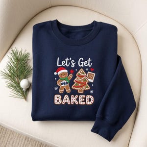 May include: A navy blue crewneck sweatshirt featuring a graphic of a gingerbread person wearing a Santa hat and holding a camera, a decorated Christmas tree, and a small photo, with the text Let's Get BAKED in white and red.