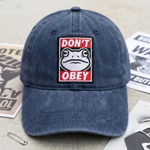 May include: A navy blue baseball cap with a curved peak. The cap has a rectangular patch with a white frog illustration and the words "DON'T OBEY" in red and white.