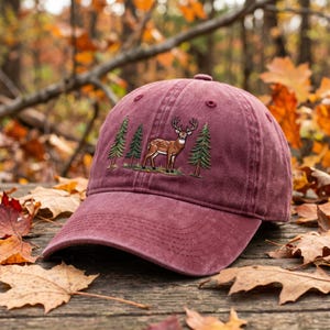 May include: A maroon baseball cap with an embroidered design of a deer and two pine trees. The cap has a curved peak and a fabric strap. The background features autumn leaves.