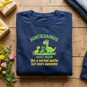 May include: A navy blue sweatshirt featuring the words "Auntiesaurus" in green, above a cartoon dinosaur family. Below are the names "Chloe | Mason" and the phrase "like a normal auntie but more awesome".