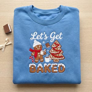 May include: A light blue crewneck sweatshirt featuring an embroidered design of a gingerbread person wearing a white hat and red scarf, holding a book. Next to it is a decorated Christmas tree, a mug, and the text Let's Get BAKED.