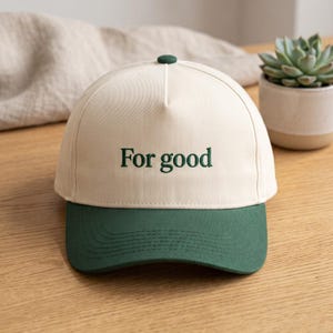 May include: A baseball cap with a cream-coloured crown and a dark green brim. The front of the crown features the text "For good" embroidered in dark green thread. The cap has a small dark green button at the top centre of the crown.