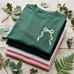 May include: A pile of folded t-shirts in shades of green, navy, white, and pink. The top green tee has a white embroidered bow and the letter "E". The shirts are stacked on a wooden surface, decorated with foliage.