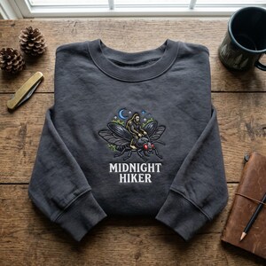 May include: A charcoal grey crewneck sweatshirt featuring an embroidered graphic of a creature riding a large insect under a crescent moon and stars, with the text MIDNIGHT HIKER printed in white capital letters below the central illustration.