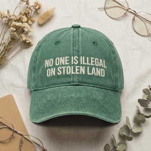 Könnte beinhalten: Eine grüne Baseballkappe mit dem gestickten Text "NO ONE IS ILLEGAL ON STOLEN LAND" in Weiß. Die Kappe hat einen gebogenen Schirm und einen Used-Look. Der Hut ist ein klassisches Sechs-Panel-Design.