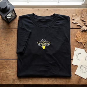 May include: A black short-sleeve t-shirt featuring a centered embroidered graphic of a bee. The bee design is composed of yellow and white thread, showcasing a detailed outline of the insect's wings and body on the solid black fabric.