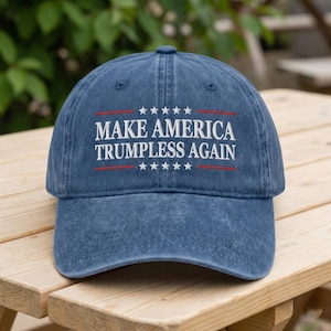 May include: A blue denim baseball cap with the text "MAKE AMERICA TRUMPLESS AGAIN" in white, with red lines and stars. The hat is sitting on a wooden surface.